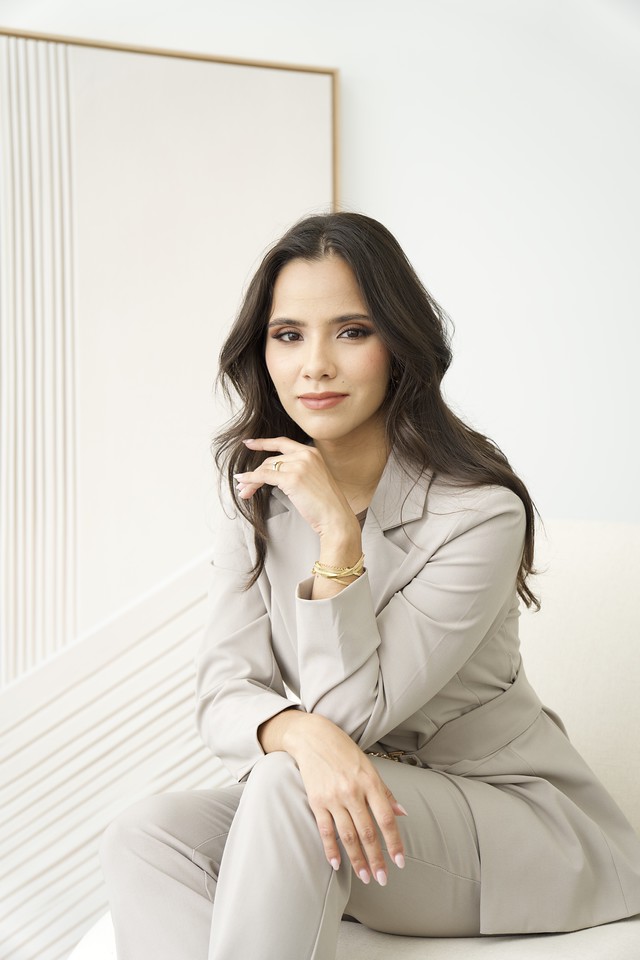 Retrato de Natália Luz, sentada, de terninho claro, em frente a um painel com frisos verticais